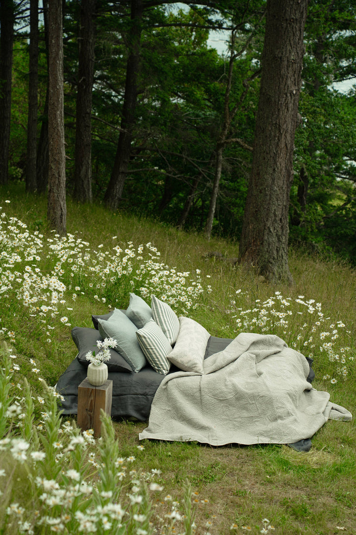 Outdoor setting with pillows and blankets on a grassy area surrounded by trees and wildflowers.