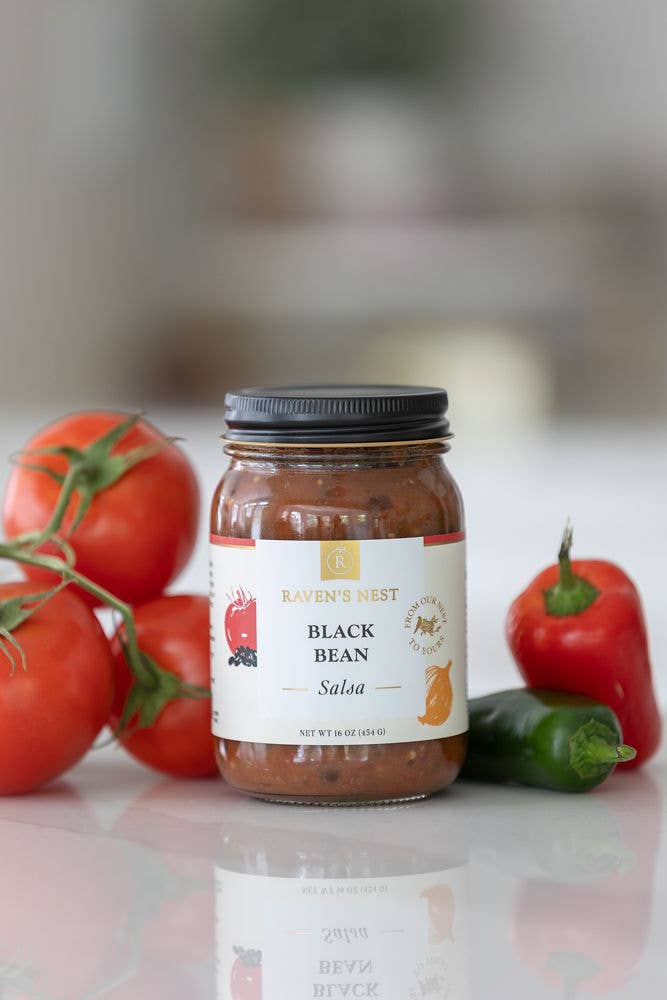 Jar of Raven's Nest Black Bean Salsa with tomatoes and a jalapeño pepper on a reflective surface.