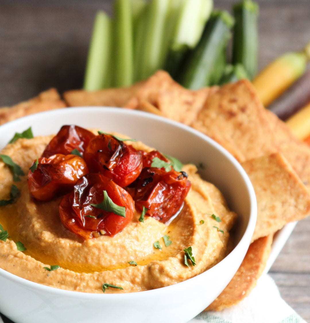 Hummus with roasted tomatoes and garden vegetable dip in a bowl with crackers and vegetables on a wooden surface