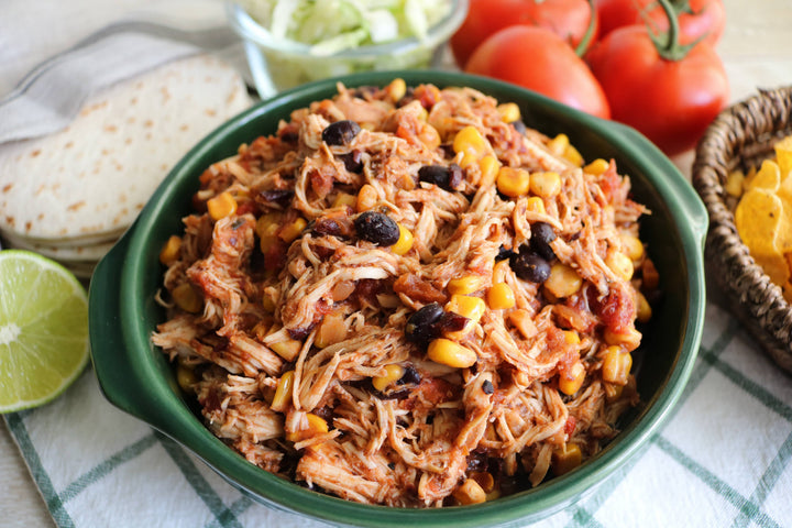 Bowl of pulled pork with corn and black beans on a table with tortillas and tomatoes.