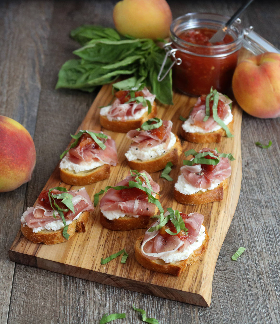 Wooden board with crostini topped with prosciutto, cheese, and basil, surrounded by peaches and a jar of jam.