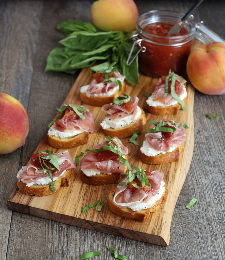 Wooden board with crostini topped with prosciutto, cheese, and basil, surrounded by peaches and a jar of jam.