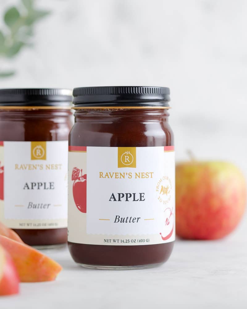 Two jars of Raven's Nest Apple Butter with apples on a light background