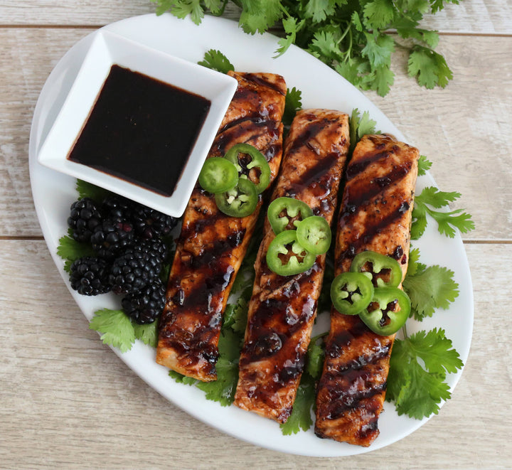 Grilled salmon with jalapeños, blackberries, and a small dish of sauce on a white plate.