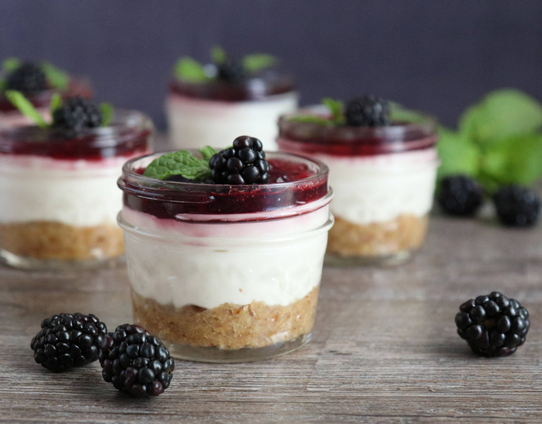 Small glass jars with layered dessert, blackberries, and mint leaves on a wooden surface.