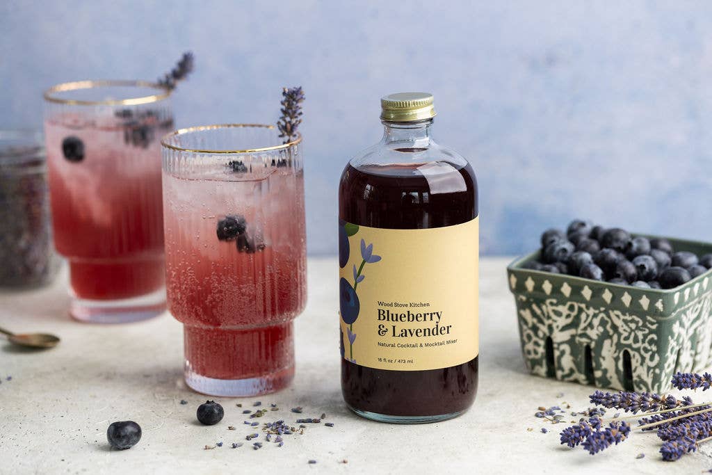 Bottle of Blueberry & Lavender syrup with two glasses of a red drink on a light surface.