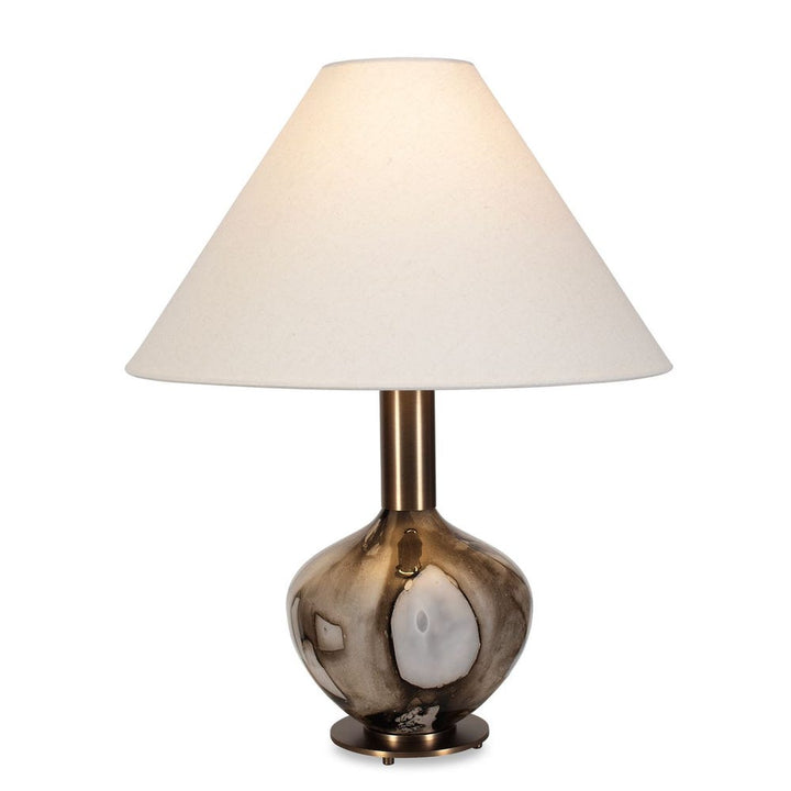 This table lamp showcases a captivating base crafted from art glass with a mesmerizing smoky, dark brown pattern. The intricate details and warm brass plated accents create a truly unique and eye-catching piece. The coolie style shade is covered in a white linen fabric. Due to the natural hand crafted art glass each piece will vary.