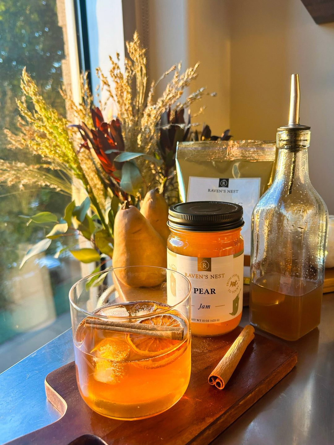 Glass of orange juice with a cinnamon stick, jar of pear jam, and bottle on a wooden board by a window.