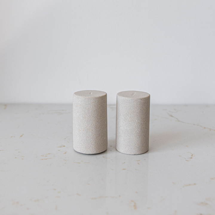 Two cylindrical stone salt and pepper shakers on a light marble surface with a white background