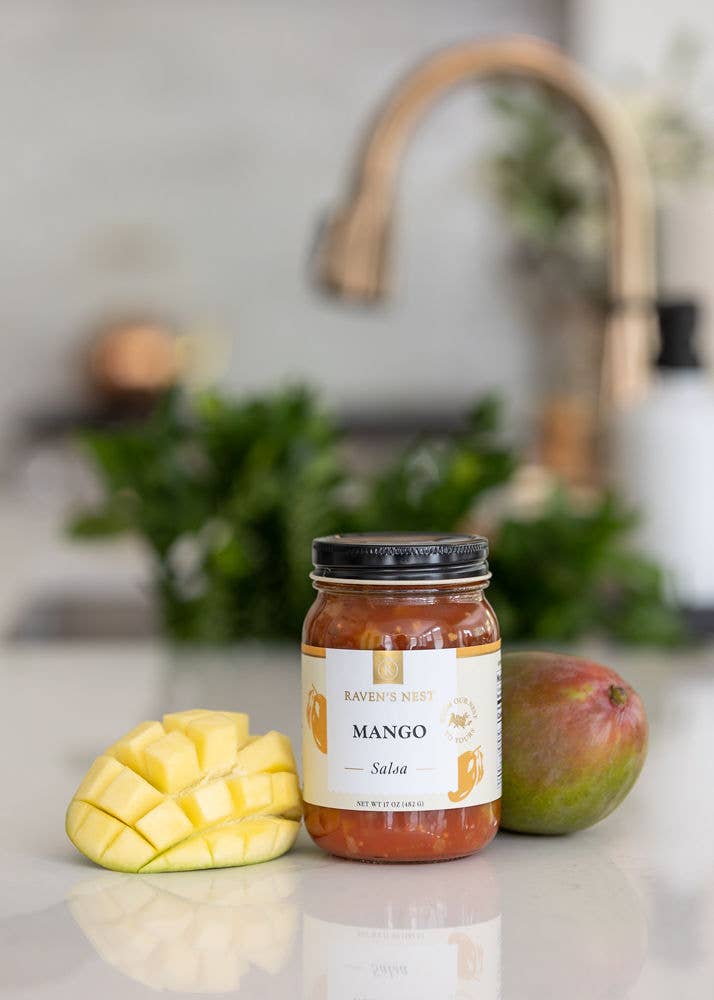Jar of mango salsa on a kitchen counter with mangoes and a blurred background