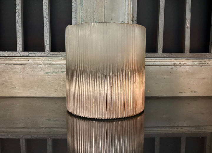 Cylindrical ribbed glass vase on a reflective surface with a wooden background