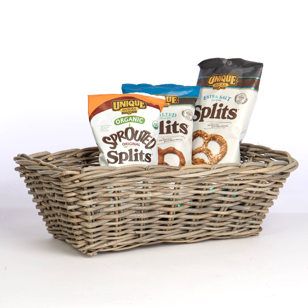 A rectangular woven wicker basket filled with packaged snack items, including bags of Unique Brands organic and seeded pretzel splits, on a clean white background.