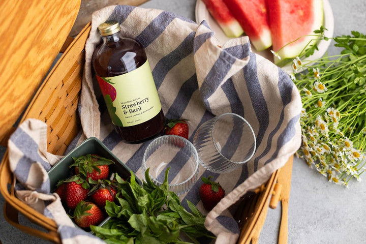 Strawberry & Basil cocktail mixer bottle on a picnic blanket with strawberries, watermelon, and flowers.