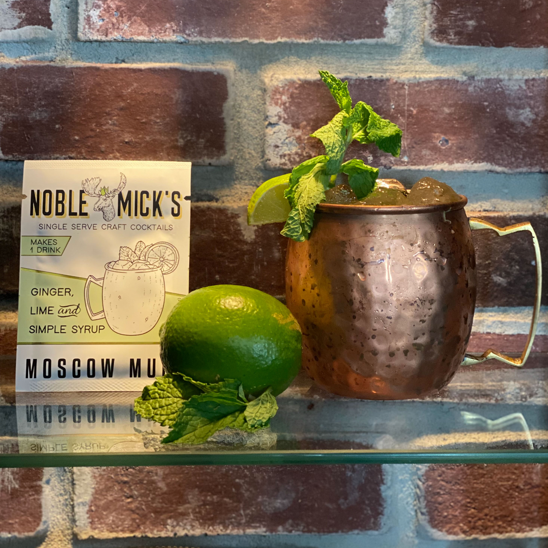 Copper mug with cocktail, lime, and mint leaves on a brick wall background with Noble Mick's Moscow Mule packaging.
