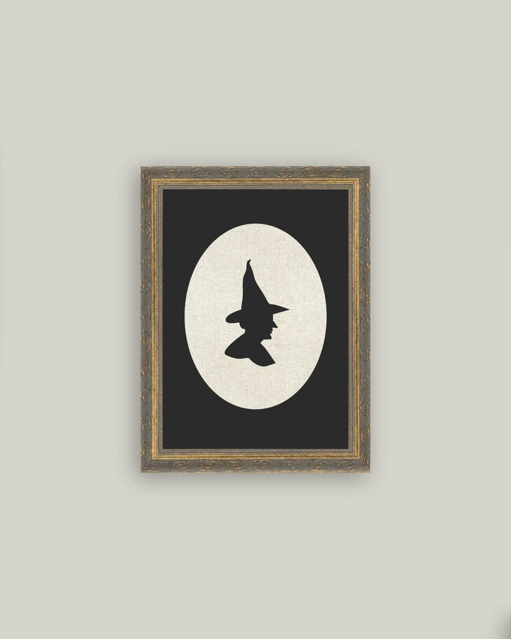 Framed artwork with a witch hat silhouette against a moonlit background on a gray wall.
