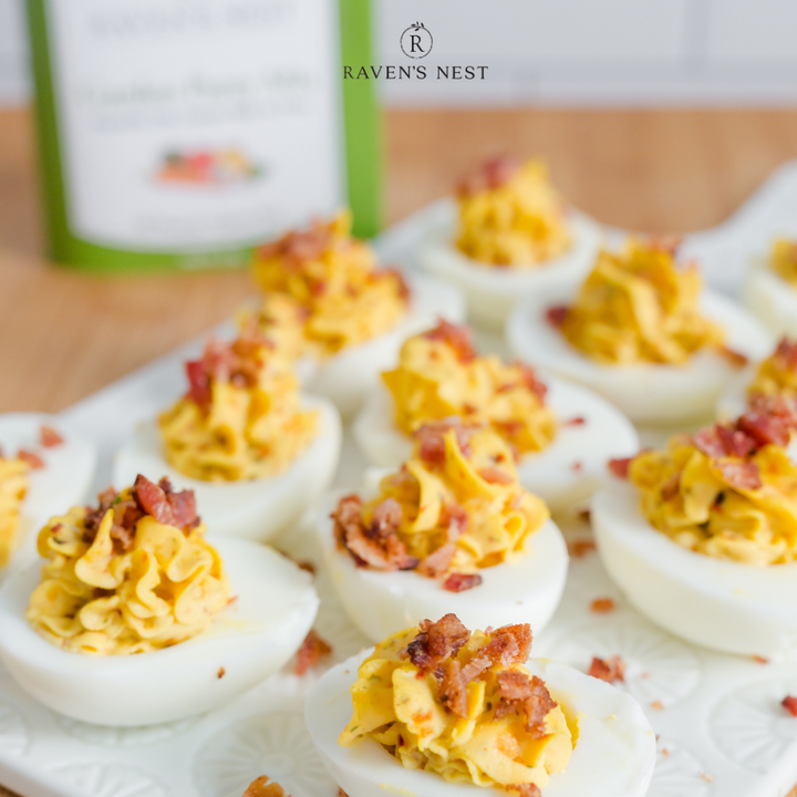Deviled eggs with bacon on a white plate, with 'Raven's Nest' branding in the background.