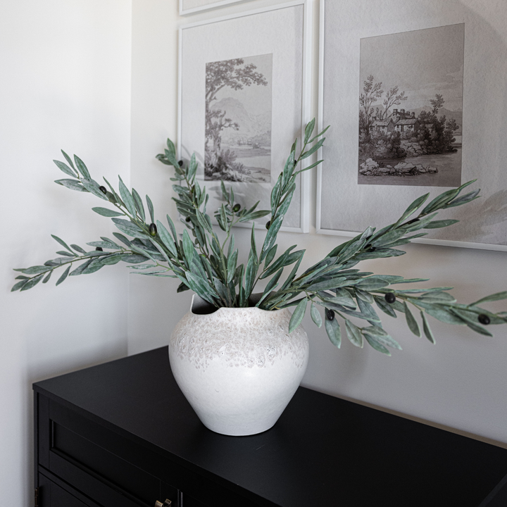 White vase with green leaves on a black cabinet against a wall with framed pictures.