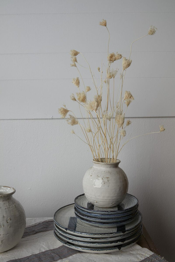 White vase with dried flowers on a stack of black plates against a light gray wall.