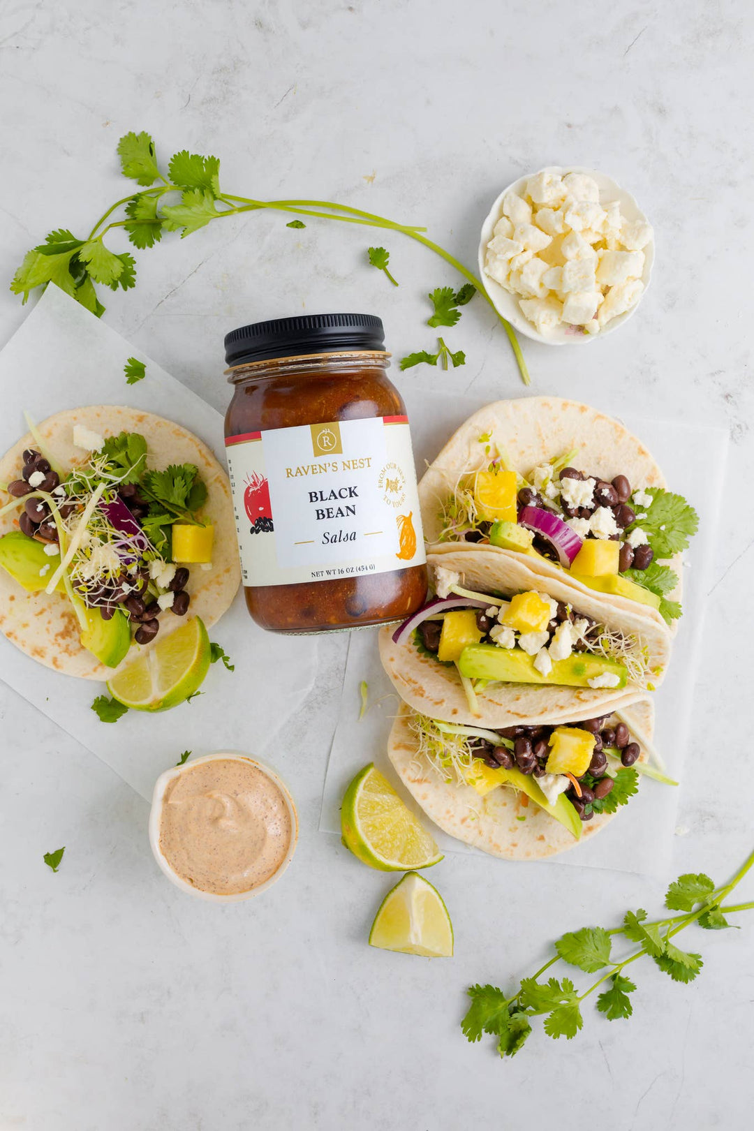 Tacos with various toppings on a white surface, featuring a jar of sauce.