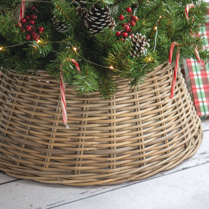 A natural woven wicker Christmas tree skirt holding green pine branches with pinecones, red berries, candy canes, and warm string lights, set on a light floor with a wrapped gift partially visible.