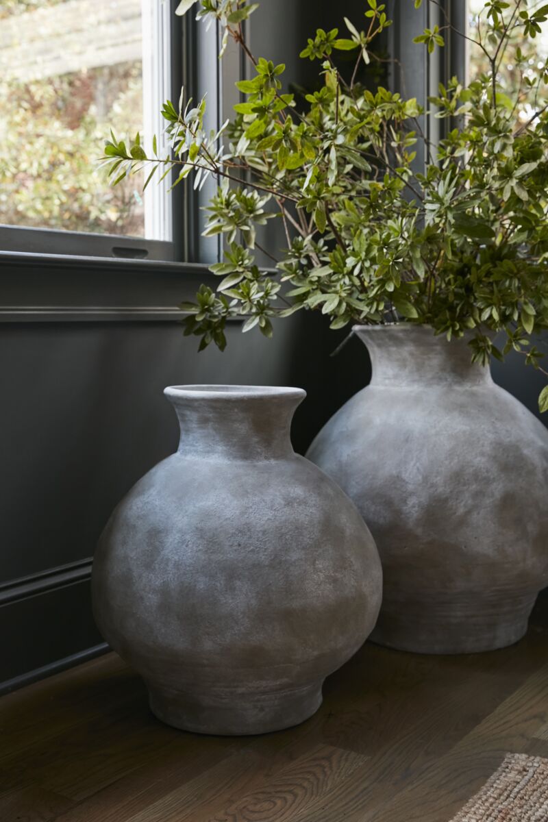 Two gray vases with greenery on a wooden surface near a window.