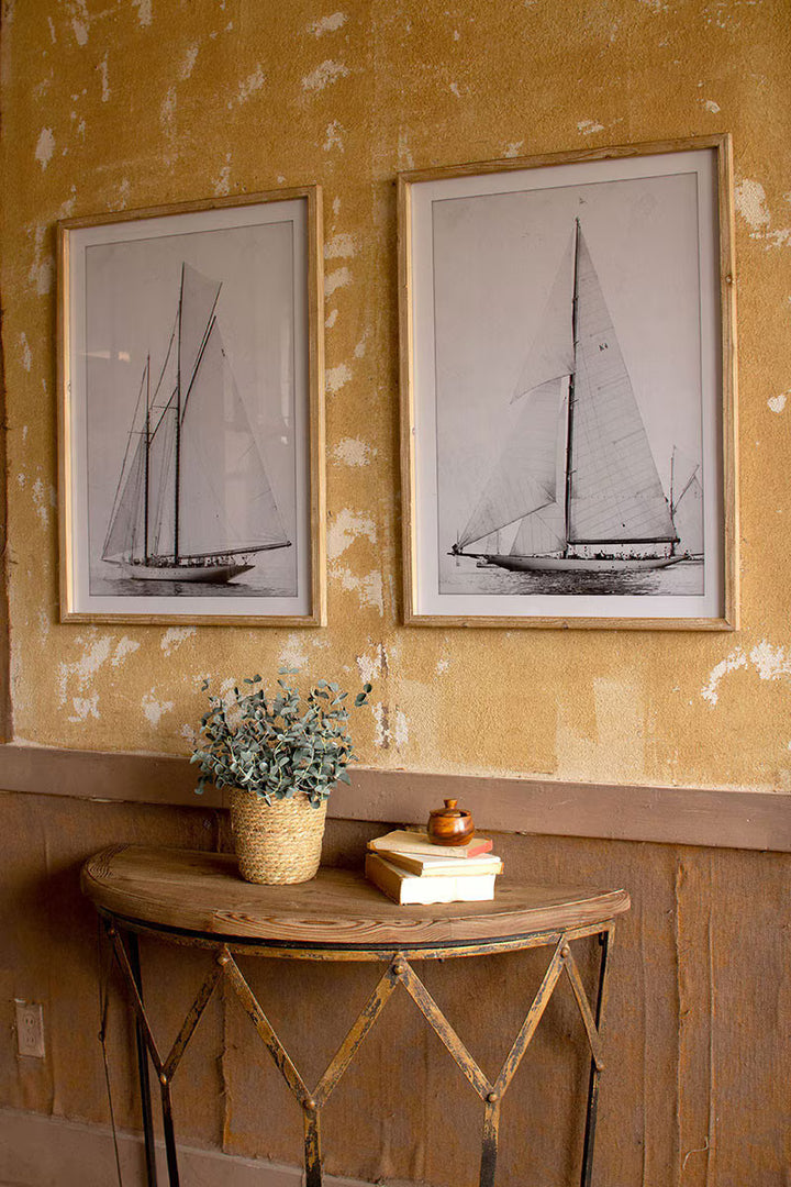 Two framed black and white sailboat prints on a textured wall with a small table 