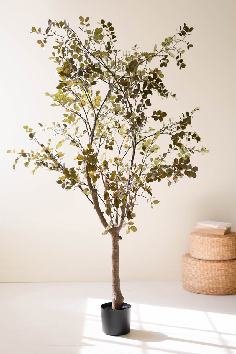 Potted artificial tree with green leaves on a white surface with woven baskets in the background.