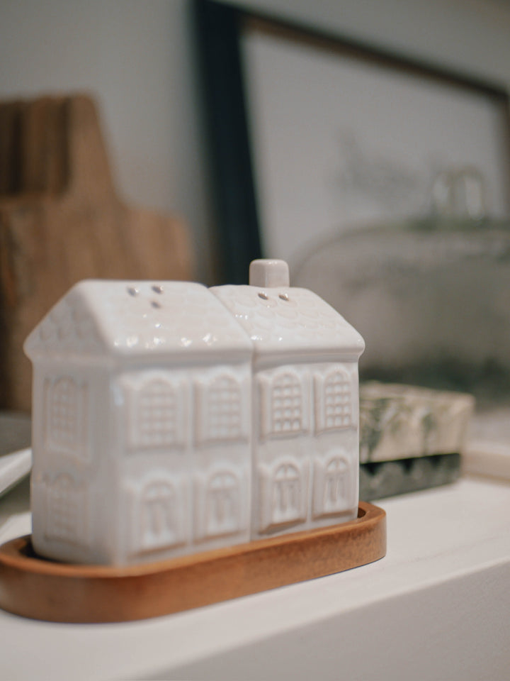 White ceramic house-shaped salt and pepper shakers on a wooden tray with a blurred background.