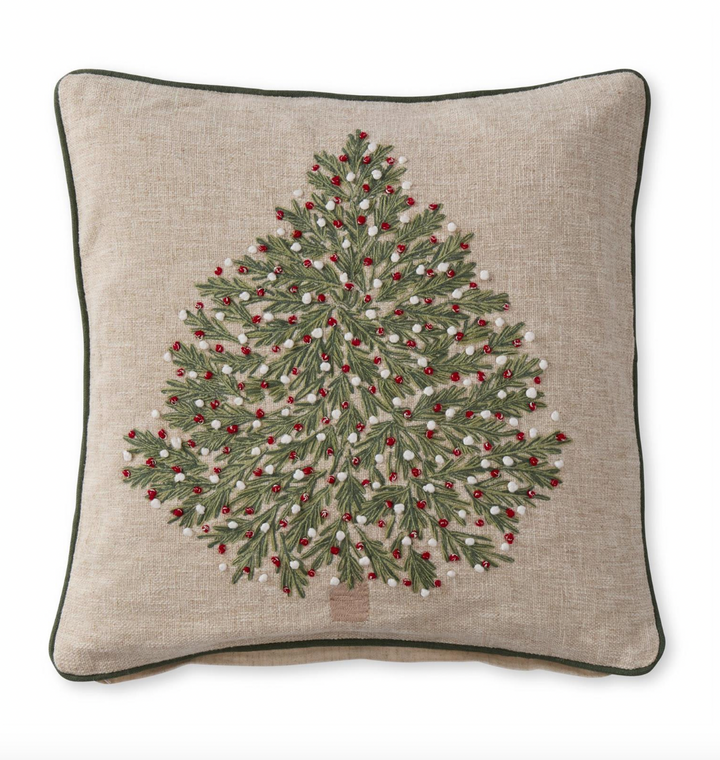 A square-shaped Christmas tree pillow with a tan background and a green Christmas tree design embellished with red and white ornaments.