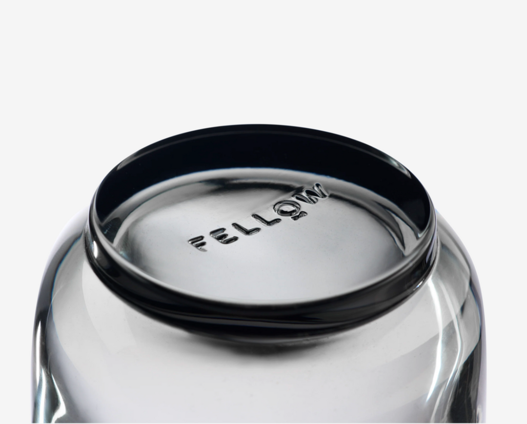 Close-up of a glass coffee mug with a black lid featuring the brand 'Fellow' on a white background.