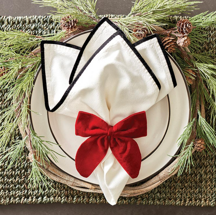 A red velvet bow napkin tie on a white napkin, presented on a plate with Christmas decor in the background.