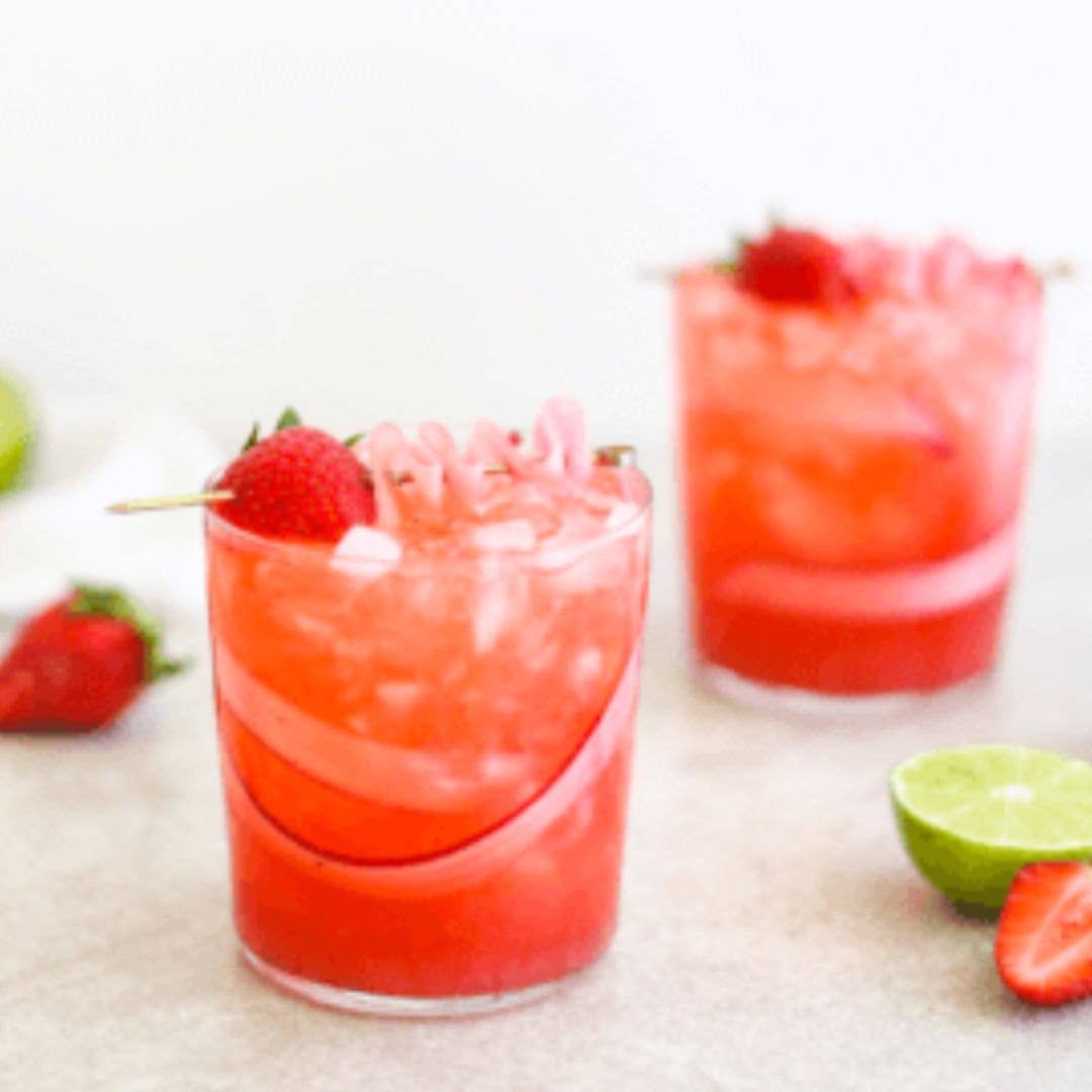 Two strawberry and basil cocktails with lime wedges and strawberries on a white surface.
