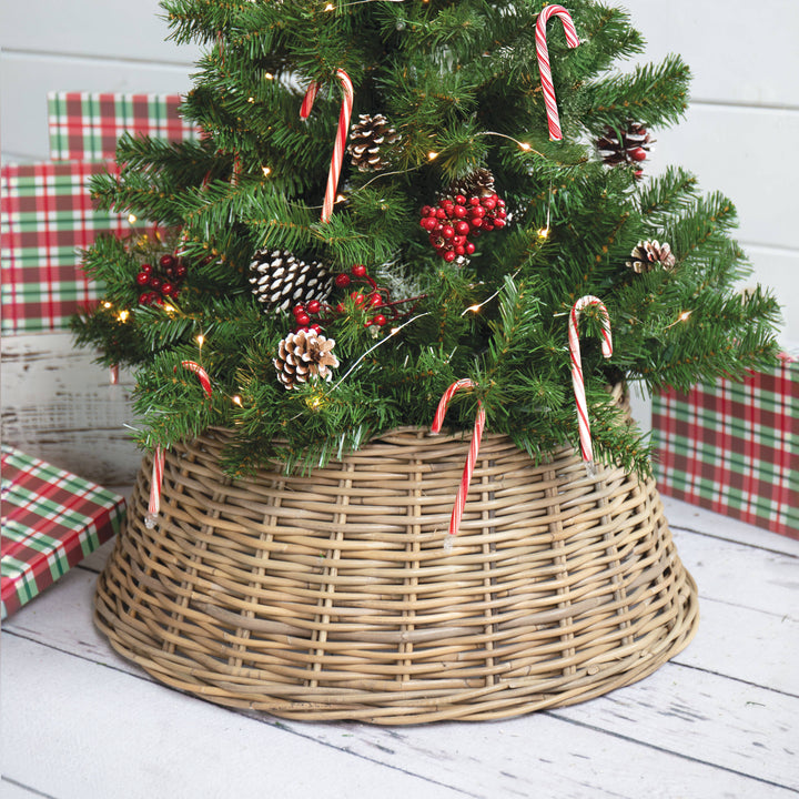 A natural wicker Christmas tree skirt holding a decorated green Christmas tree with candy canes, pinecones, and red berries, styled with wrapped presents on a white wooden floor.