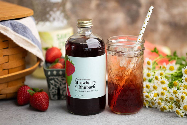 Bottle of strawberry rhubarb syrup next to a glass of iced tea with strawberries and flowers in the background.
