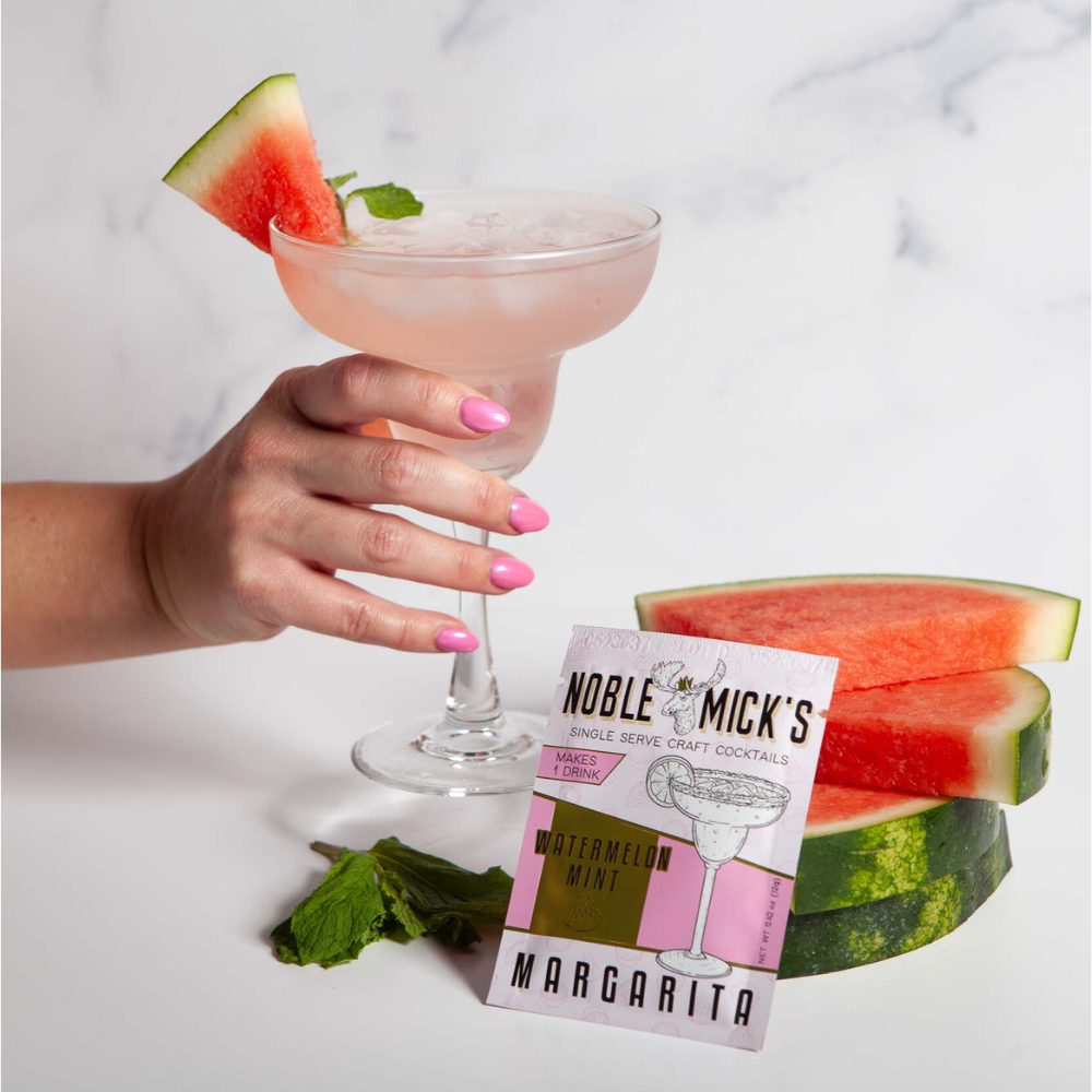 Hand holding a cocktail with watermelon garnish, next to watermelon slices and a cocktail package on a light background