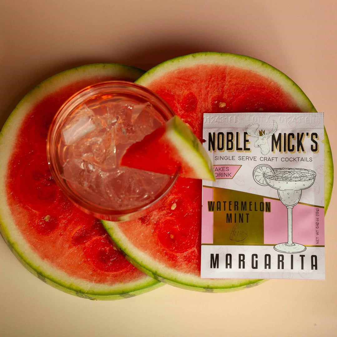 Glass of pink cocktail with ice cubes on watermelon slices next to a 'Noble Mick's' cocktail package.