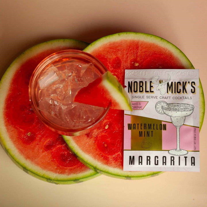 Glass of pink cocktail with ice cubes on watermelon slices next to a 'Noble Mick's' cocktail package.
