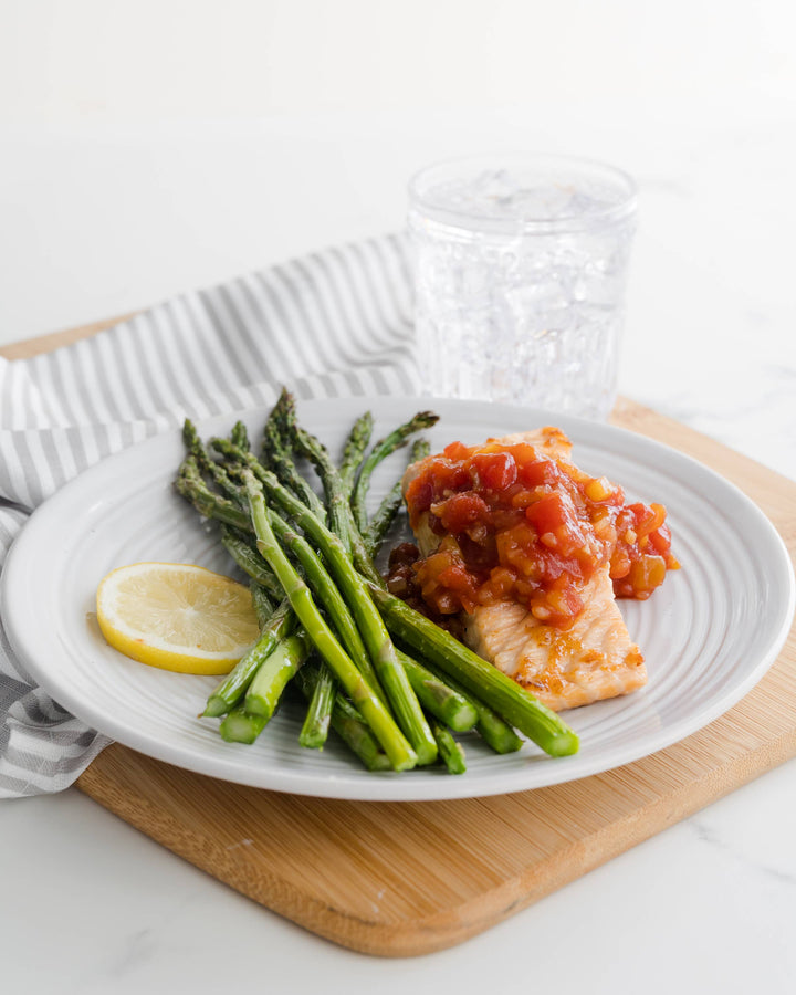 Plate of salmon with tomato salsa and asparagus on a white plate with a lemon wedge, on a wooden board.