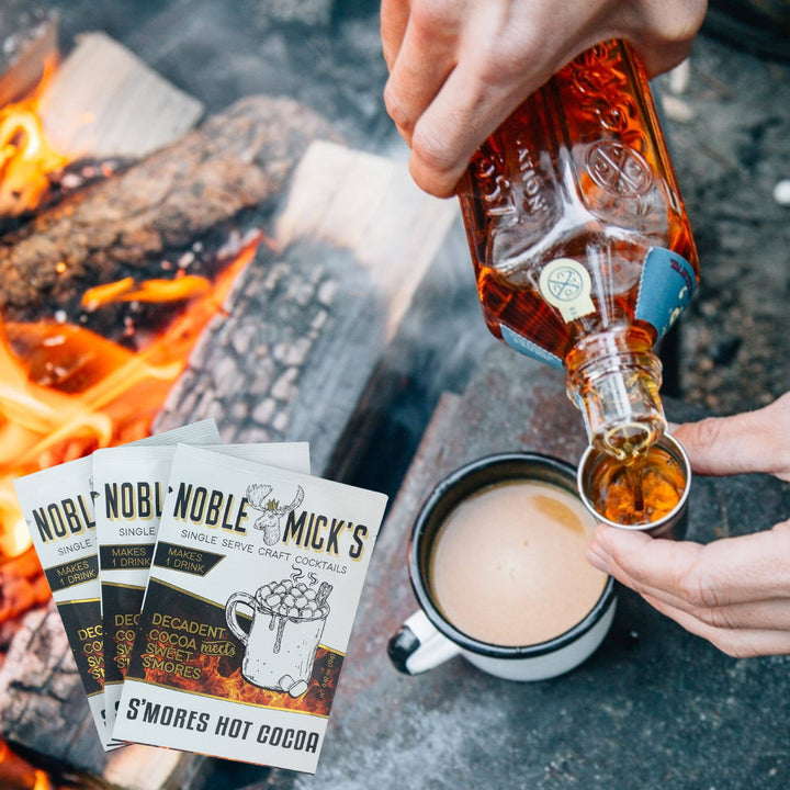 Person pouring a liquid into a mug next to 'Noble Mick's' cocktail packages by a campfire.