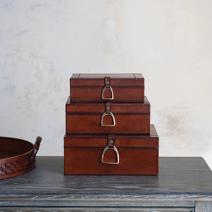Three stacked brown leather boxes with metal handles on a wooden surface.