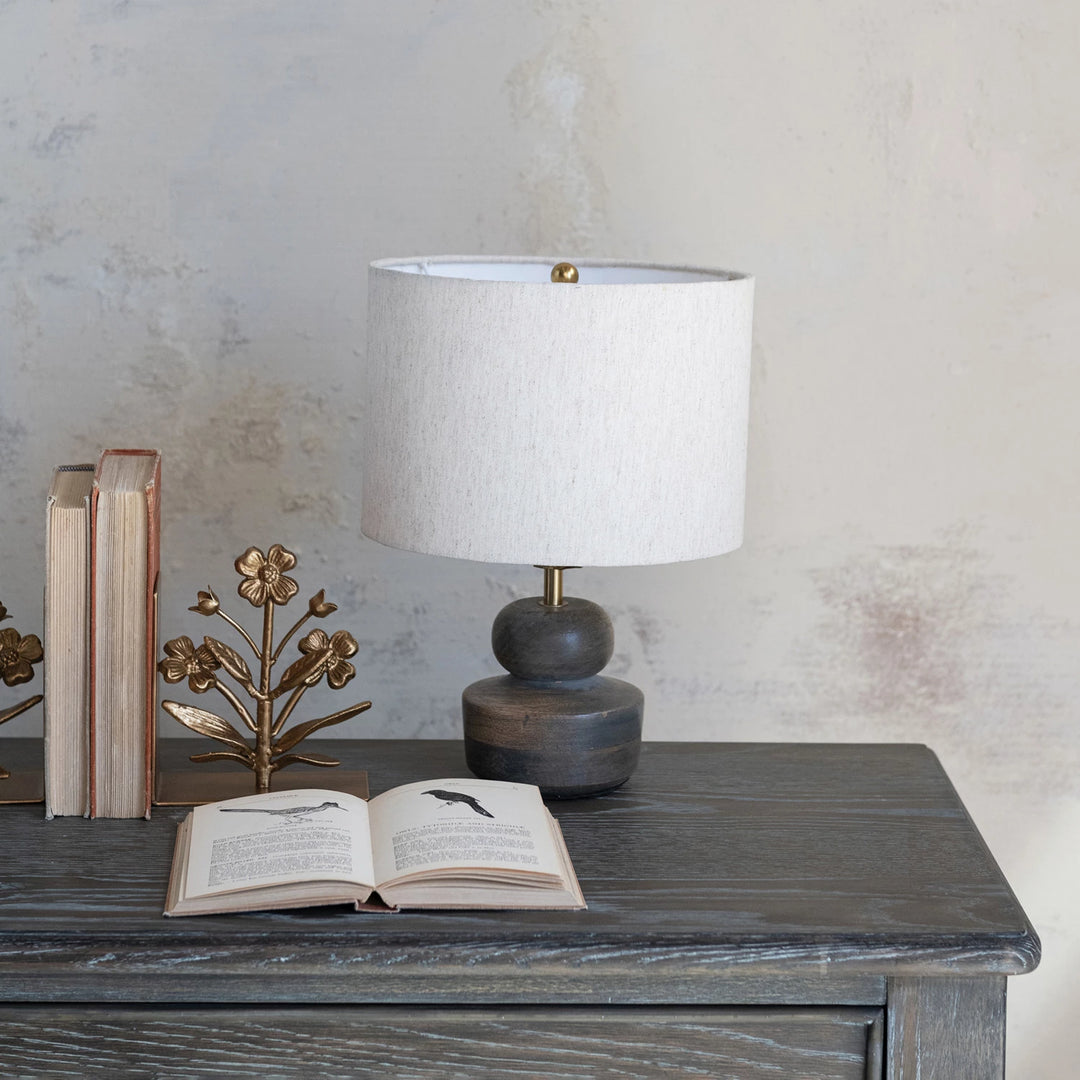 Table lamp with white shade on a wooden surface next to an open book and decorative items.