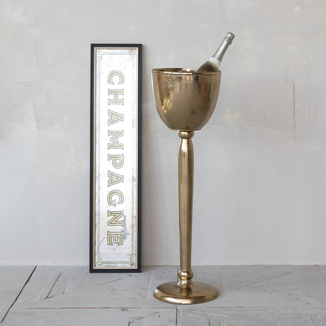 Gold champagne bucket with bottle against a white wall with 'Champagne' sign.