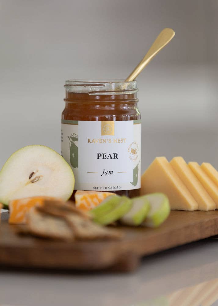 Jar of pear jam with a spoon, apples, and cheese on a wooden board.