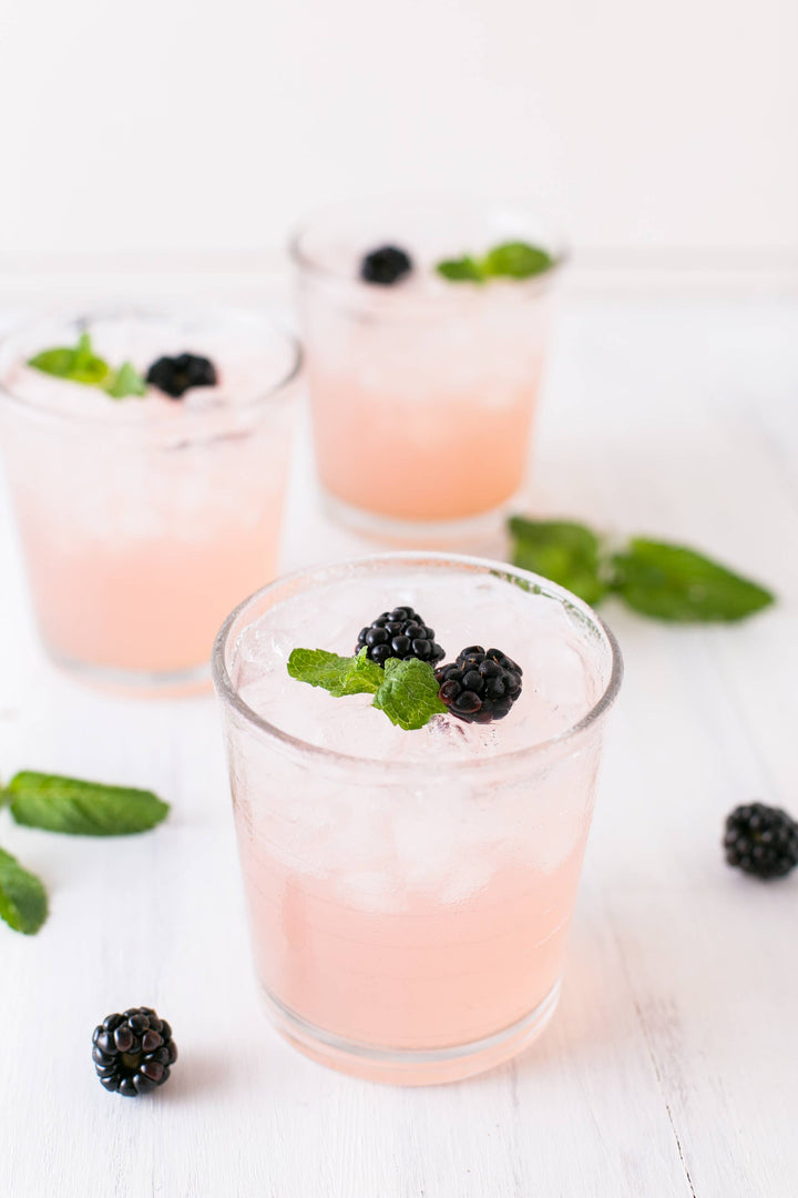Three pink drinks in glasses with blackberries and mint leaves on a white surface.