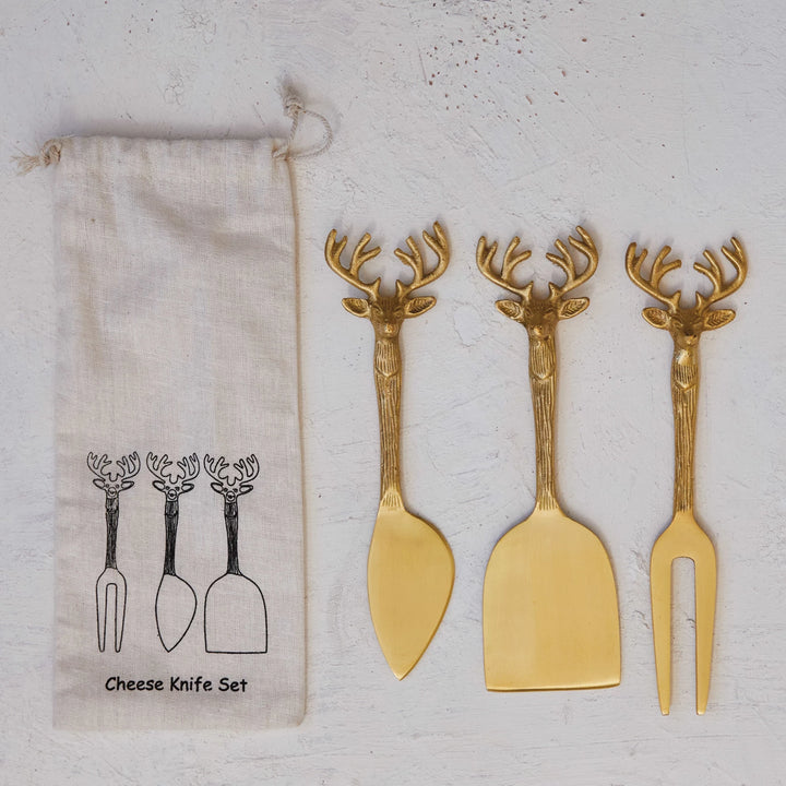 A set of three brass cheese knives with reindeer handles, displayed alongside a printed drawstring bag 