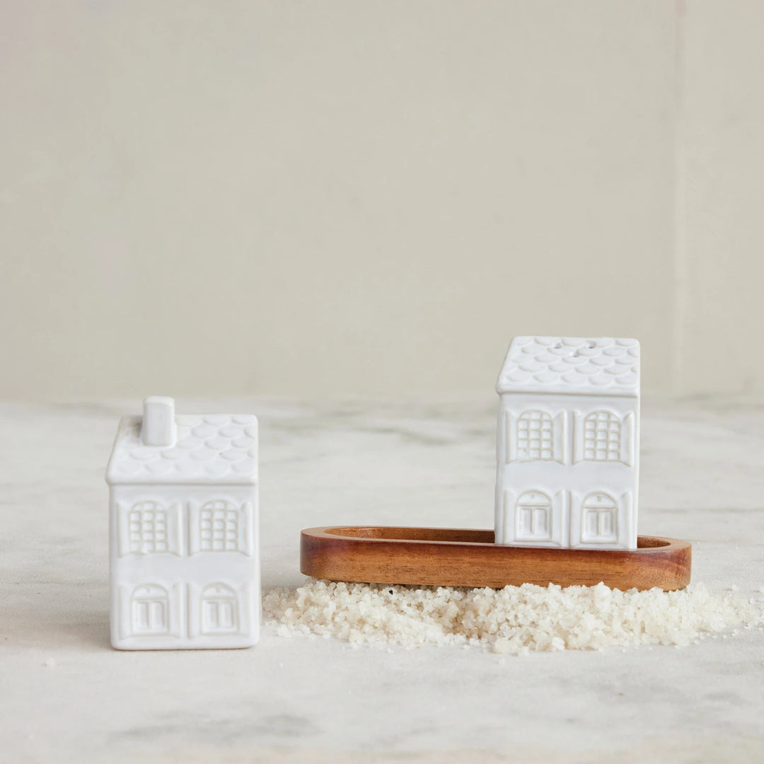 A set of white stoneware salt and pepper shakers designed to look like houses, displayed on a wooden tray filled with salt.